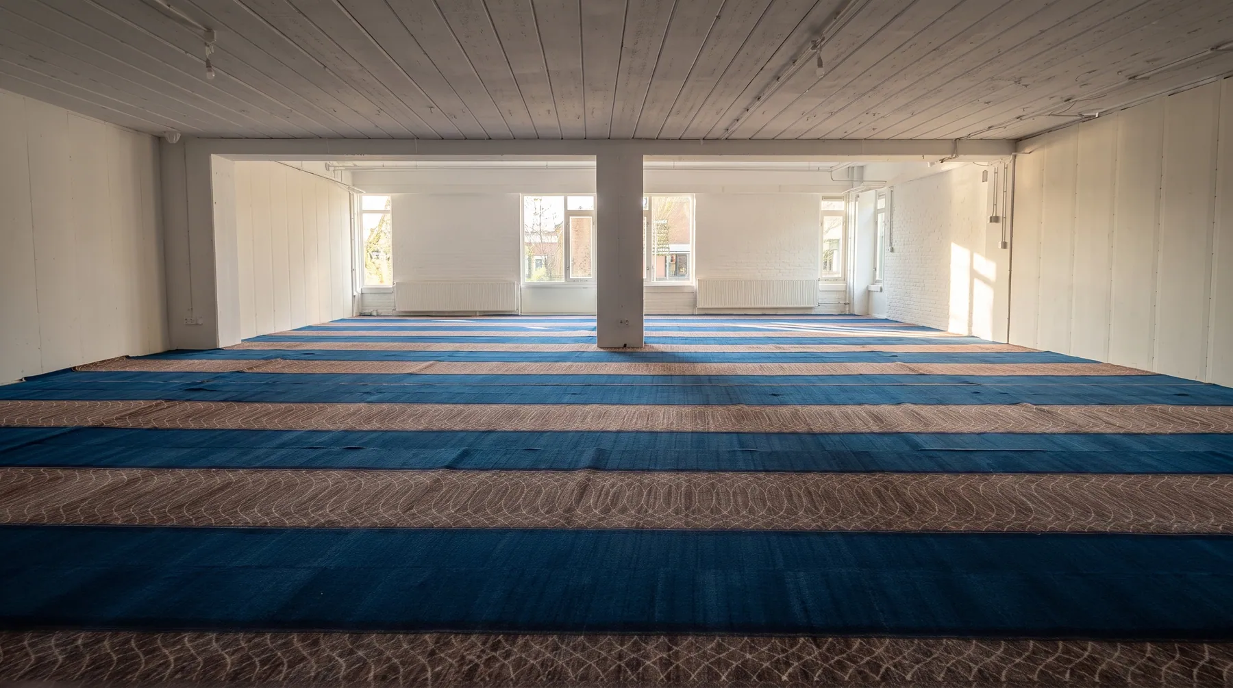 Rijen gebedstapijt in blauw en zand vullen de lichte, ruime gebedszaal